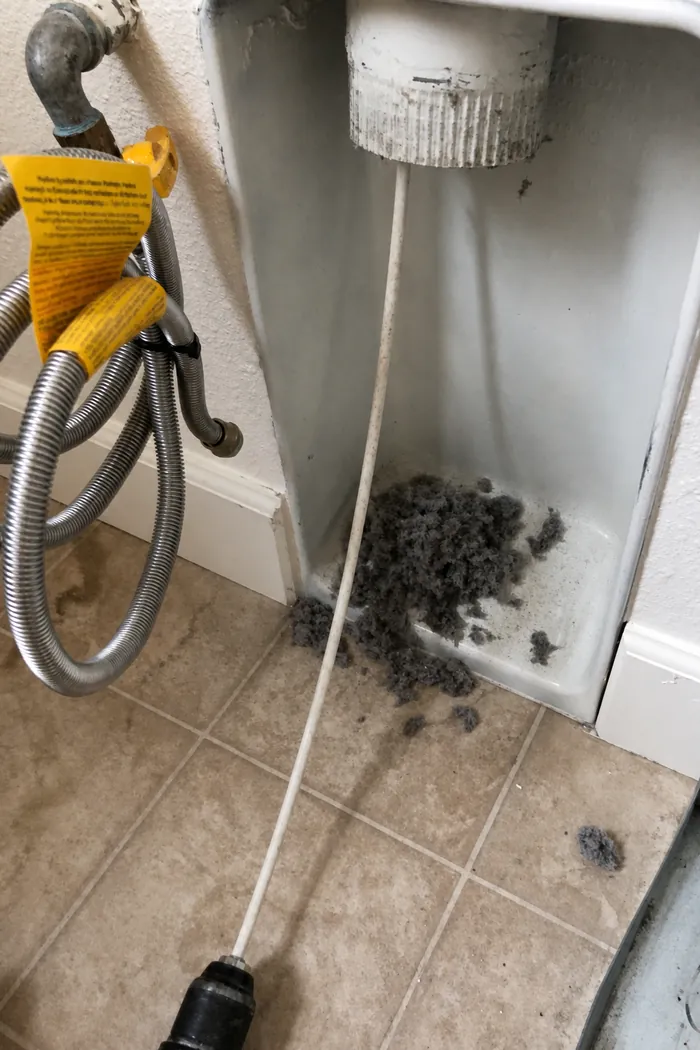 A drill-driven flexible rod cleaning a wall vent box, lint falling to the floor